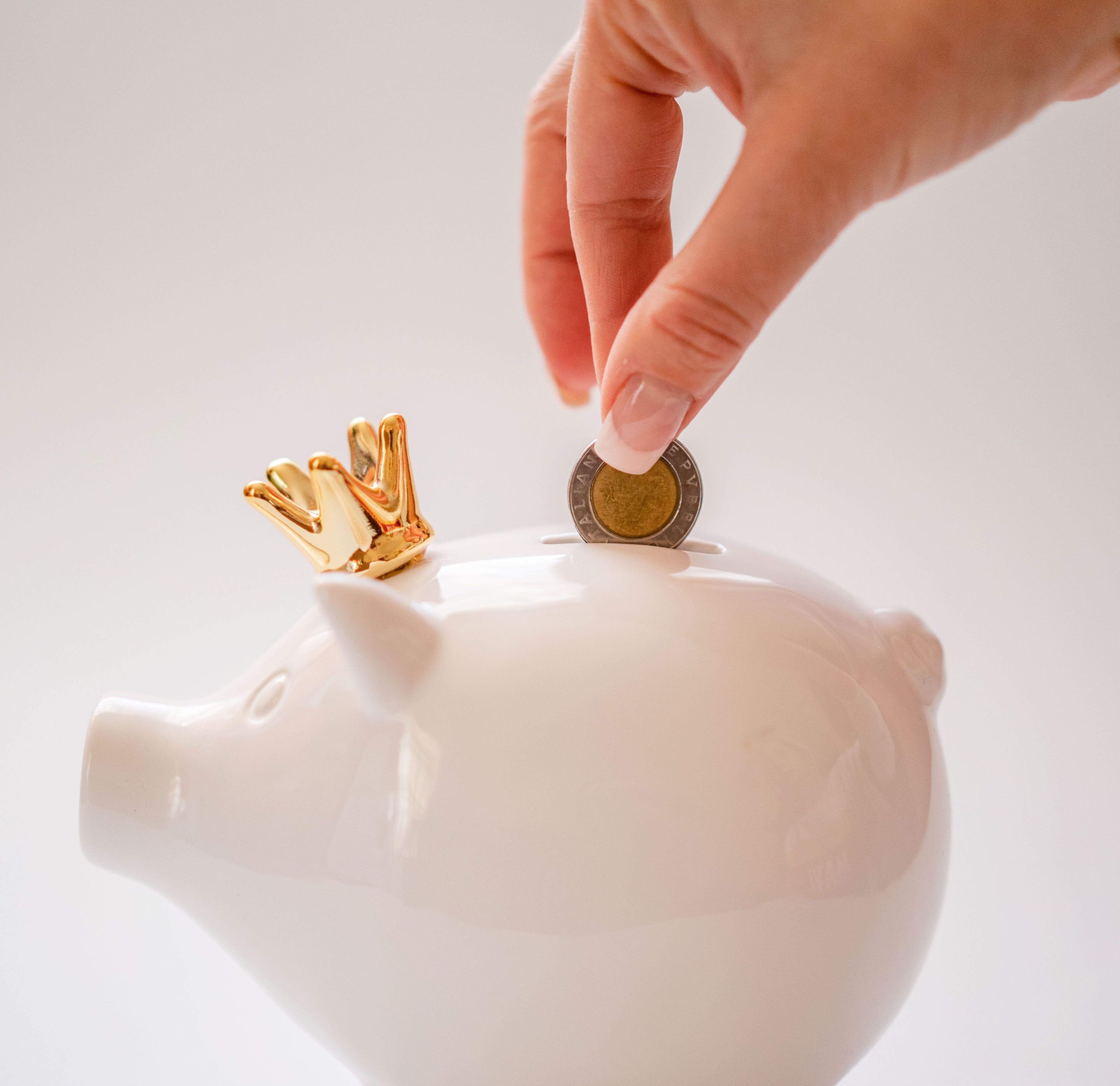 Mão depositando uma moeda em um cofrinho branco com coroa dourada
